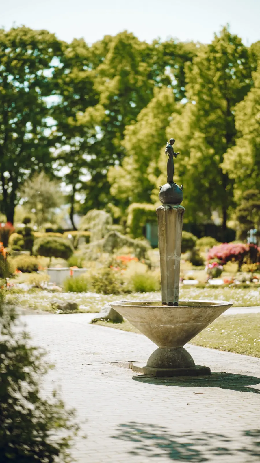 Sonniger Friedhof mit altem Brunnen und blühenden Beeten